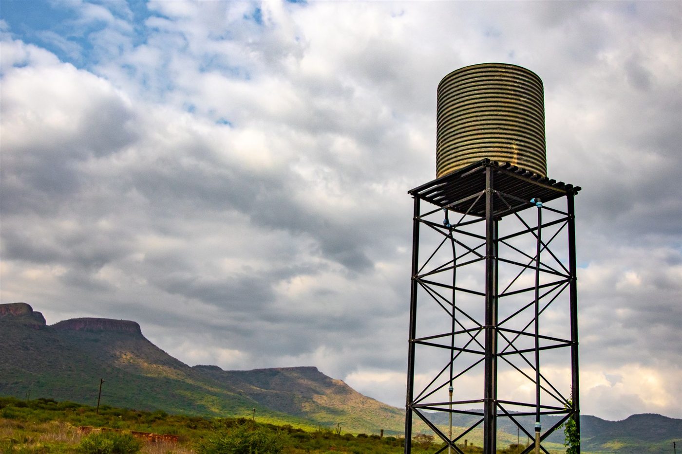 Steel water tower carrying a storage tank against a cloudy sky, with hills in the distance