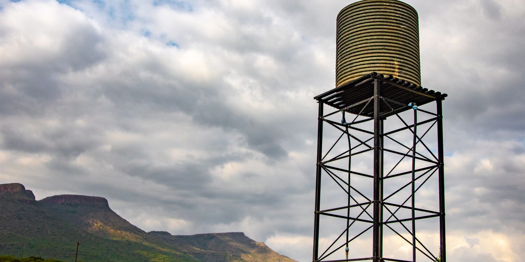Steel water tower carrying a storage tank against a cloudy sky, with hills in the distance